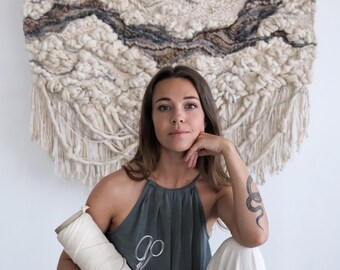 May include: A large, textured wall hanging in cream, beige, and brown tones hangs behind a person. The person is wearing a gray halter top with the word "go" and scissors printed on it, and holding a spool of white thread and scissors. The person is also wearing white pants.