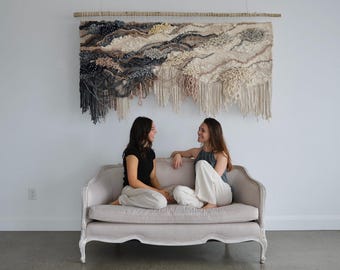 May include: Two people sitting on a white couch in front of a large woven wall hanging. The wall hanging is made of different shades of brown, beige, and gray yarn. The couch is a light gray color with a white fabric covering.