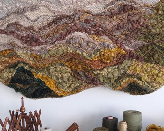 May include: A large, textured wall hanging with a landscape design in shades of beige, brown, green, and yellow. Below, a wooden table displays yarn, spools, and tools for fiber art, including a swift and a ball winder.
