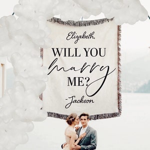 May include: A couple is standing in front of a white banner with the text "Elizabeth, WILL YOU marry ME? - Jackson". The banner is decorated with white balloons and is hanging from a white archway. The couple is standing on a balcony with a wrought iron railing.