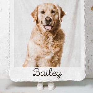 May include: A white blanket featuring a golden retriever dog with the name "Bailey" in cursive script at the bottom. The dog is light brown with its tongue out. The blanket is hanging against a white brick wall.