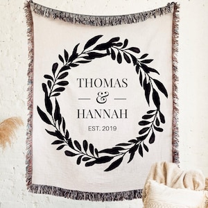 May include: Cream-colored woven throw blanket with black fringe and a black wreath design. The wreath surrounds the names "THOMAS & HANNAH" and the year "EST. 2019". The blanket is hanging on a white brick wall.