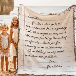 K&ouml;nnte beinhalten: Eine cremefarbene Decke mit einem handgeschriebenen Brief an "Mama" von "Euren Kindern". Der Text ist in schwarzer Tinte, die an Notizpapier erinnert. Drei junge M&auml;dchen stehen an einem Sandstrand und halten die Decke. Der Ozean und eine Felsklippe sind im Hintergrund.