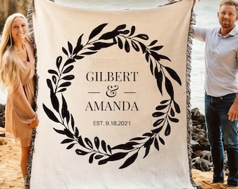 May include: Cream-colored throw blanket with a black leaf wreath design. The blanket features the names "Gilbert & Amanda" and the date "EST. 9.18.2021". The blanket is fringed and held up on a beach.