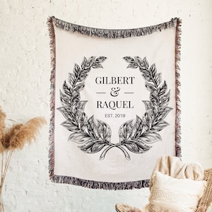 May include: A white throw blanket with a black and white wreath design. The wreath has the text "Gilbert & Raquel Est. 2019" in the center. The blanket is hanging on a white brick wall.