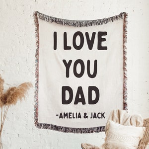 May include: A white woven tapestry with black text that reads "I love you Dad - Amelia & Jack". The tapestry is hanging on a white brick wall. A wicker chair with a white pillow and a brown fur throw is in the foreground.