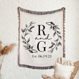 May include: A woven throw blanket with a cream background and black fringe. The blanket features a black and white monogram with the letters "R and G" surrounded by a wreath of leaves. The text "Est. 06.19.23" is printed below the monogram.