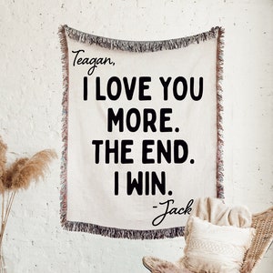 May include: A white throw blanket with black text that reads "Teagan, I love you more. The end. I win. -Jack". The blanket is hanging on a white brick wall with a wicker chair and a throw pillow in the foreground.