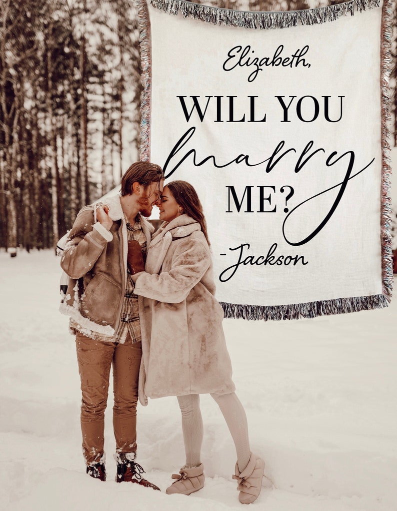 May include: A couple in a snowy forest with a white blanket behind them that says "Elizabeth, WILL YOU marry ME? - Jackson".