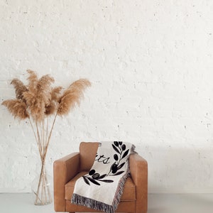 May include: A brown leather armchair with a woven throw blanket featuring a black and white olive branch design and the text "Live Simply". The chair is in front of a white brick wall with a vase of dried pampas grass.