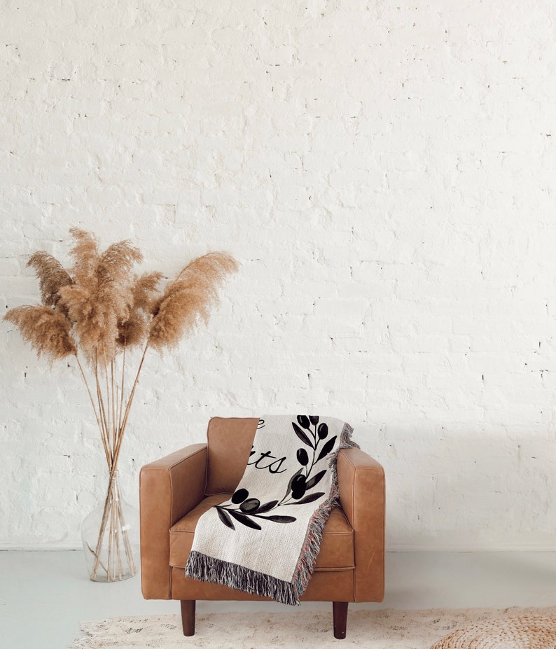 May include: A brown leather armchair with a woven throw blanket featuring a black and white olive branch design and the text "Live Simply". The chair is in front of a white brick wall with a vase of dried pampas grass.
