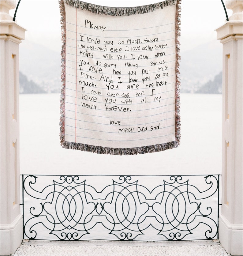 K&ouml;nnte beinhalten: Eine cremefarbene Decke mit handschriftlichem Briefdesign. Der Text lautet "Mama, ich liebe dich so sehr...love, Mitch und Syd." Die Decke hat einen Fransenrand und h&auml;ngt im Freien.