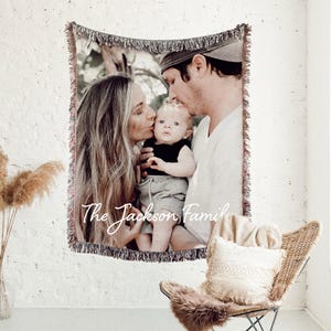 May include: A woven tapestry blanket featuring a family portrait with the text "The Jackson Family". The blanket has a fringed edge and is hanging on a white brick wall. A wicker chair with a cushion and fur throw is in the foreground.
