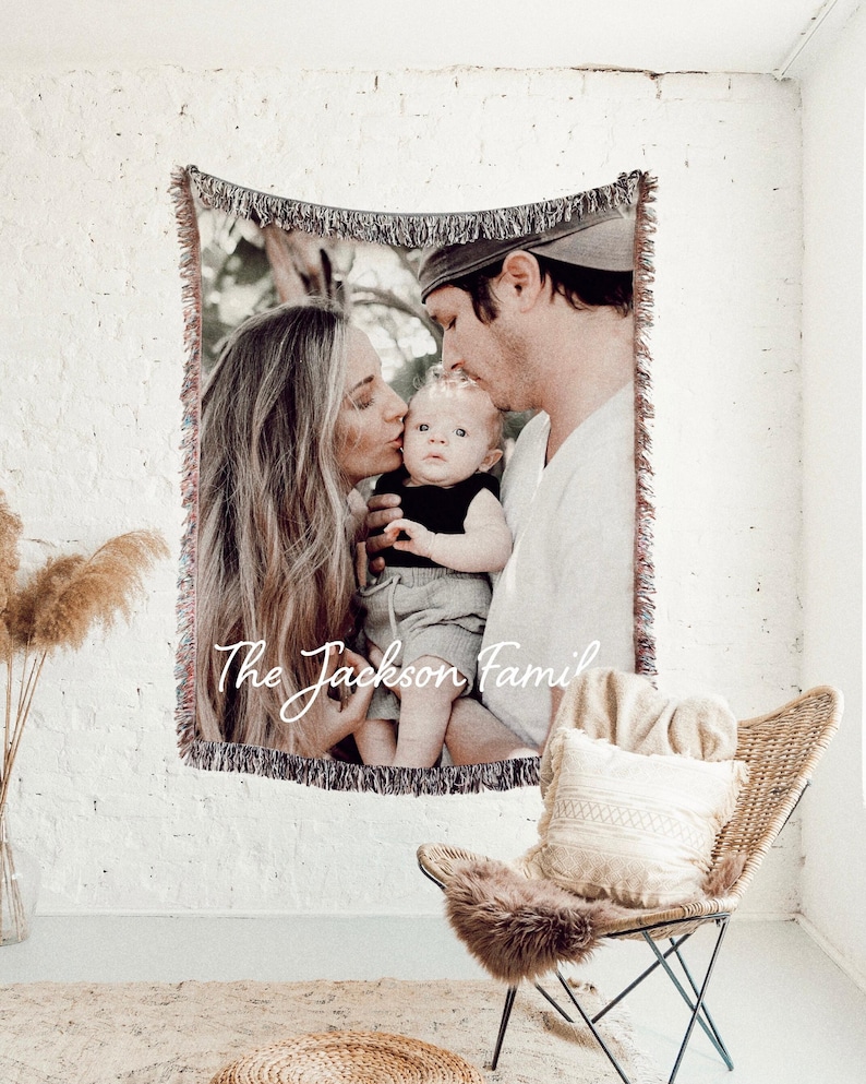 May include: A woven tapestry blanket featuring a family portrait with the text "The Jackson Family". The blanket has a fringed edge and is hanging on a white brick wall. A wicker chair with a cushion and fur throw is in the foreground.