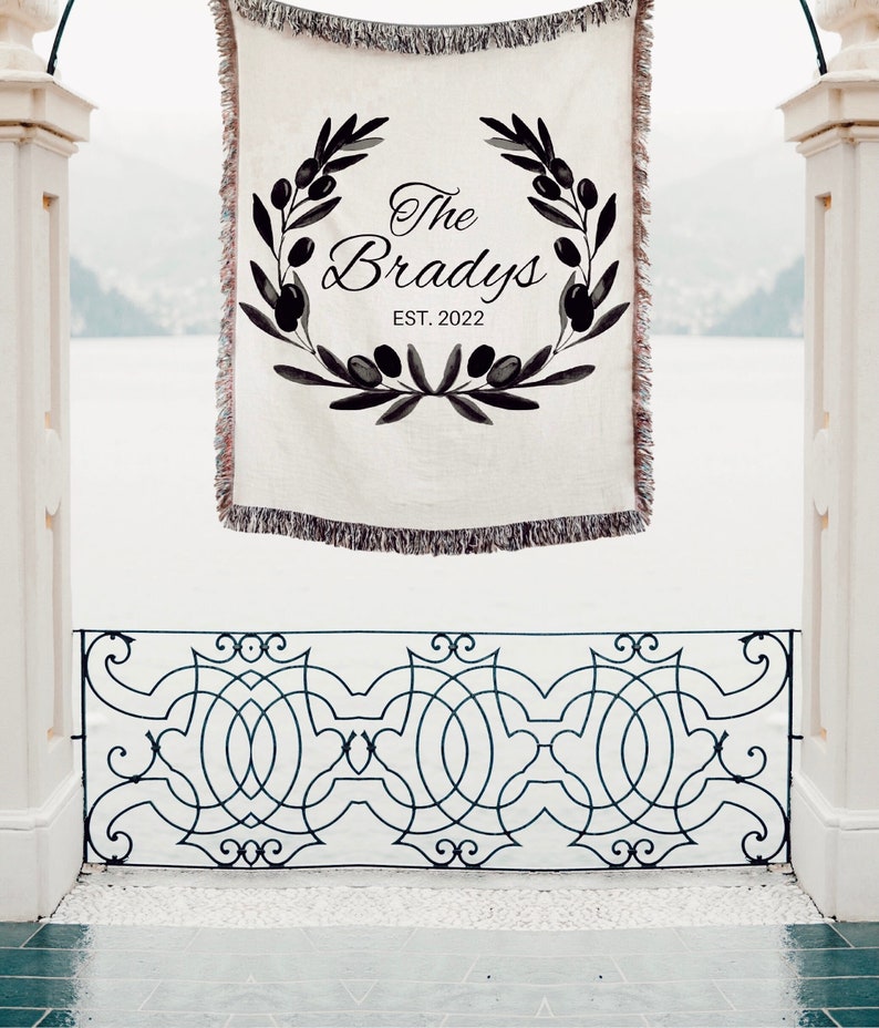 May include: A white woven throw blanket with black fringe. The blanket features a black and white illustration of an olive branch wreath with the text "The Bradys EST. 2022".