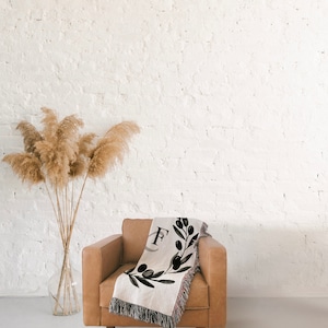 Peut inclure: Un fauteuil en cuir marron avec une couverture en laine tiss&eacute;e orn&eacute;e d'une branche d'olivier noire et blanche et de la lettre "F" au centre. La couverture est jet&eacute;e sur le dossier du fauteuil.