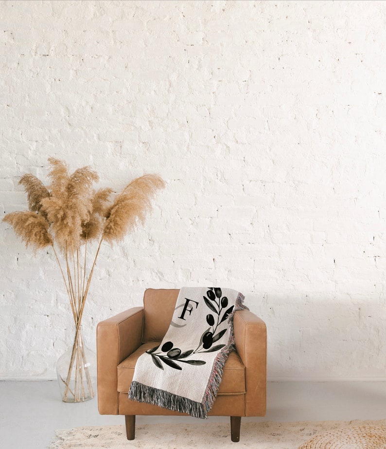 Peut inclure: Un fauteuil en cuir marron avec une couverture en laine tiss&eacute;e orn&eacute;e d'une branche d'olivier noire et blanche et de la lettre "F" au centre. La couverture est jet&eacute;e sur le dossier du fauteuil.