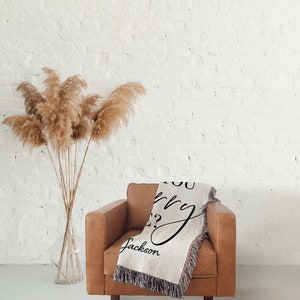 May include: A woven throw blanket with the text "Do you worry?" and the name "Jackson" in cursive script. The blanket is draped over a brown leather armchair in front of a white brick wall.