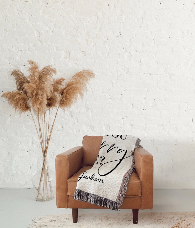 May include: A woven throw blanket with the text "Do you worry?" and the name "Jackson" in cursive script. The blanket is draped over a brown leather armchair in front of a white brick wall.