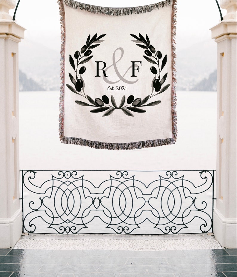 Peut inclure: Une tapisserie tiss&eacute;e blanche avec des motifs de branches d'olivier noirs et les initiales "R&F" dans un cercle gris avec le texte "Est. 2021" en dessous. La tapisserie est suspendue &agrave; une arche.