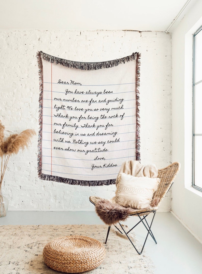 K&ouml;nnte beinhalten: Eine gewebte Decke in Creme mit einem handgeschriebenen Briefdesign, das "Liebe Mama" liest und Dankbarkeit ausdr&uuml;ckt. Die Decke hat blaue Linien und schwarzen Fransen. Sie h&auml;ngt an einer wei&szlig;en Backsteinmauer.