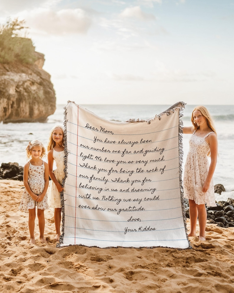 K&ouml;nnte beinhalten: Eine cremefarbene Decke mit einem handgeschriebenen Brief an "Mama" von "Euren Kindern". Der Text ist in schwarzer Tinte, die an Notizpapier erinnert. Drei junge M&auml;dchen stehen an einem Sandstrand und halten die Decke. Der Ozean und eine Felsklippe sind im Hintergrund.