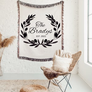 May include: A woven throw blanket with a black and white design featuring a wreath of olive branches and the text "The Bradys EST. 2022".