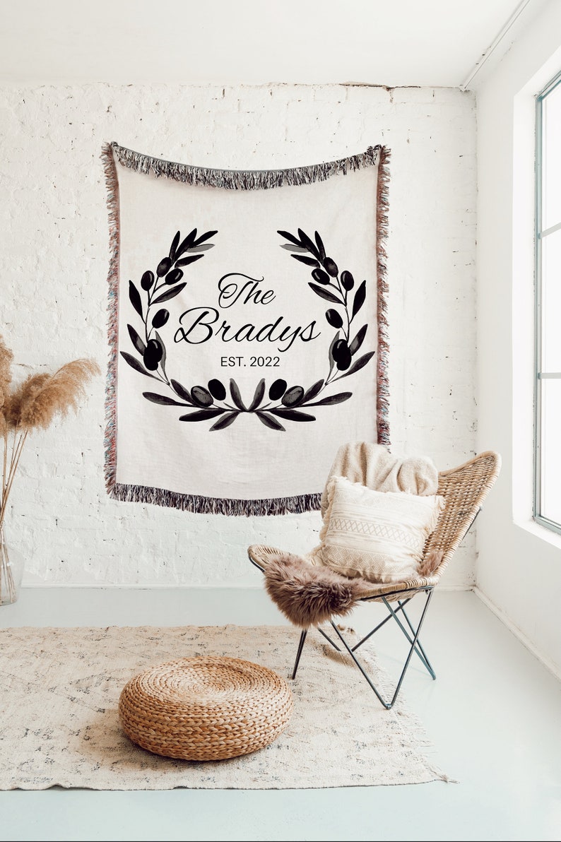 May include: A woven throw blanket with a black and white design featuring a wreath of olive branches and the text "The Bradys EST. 2022".