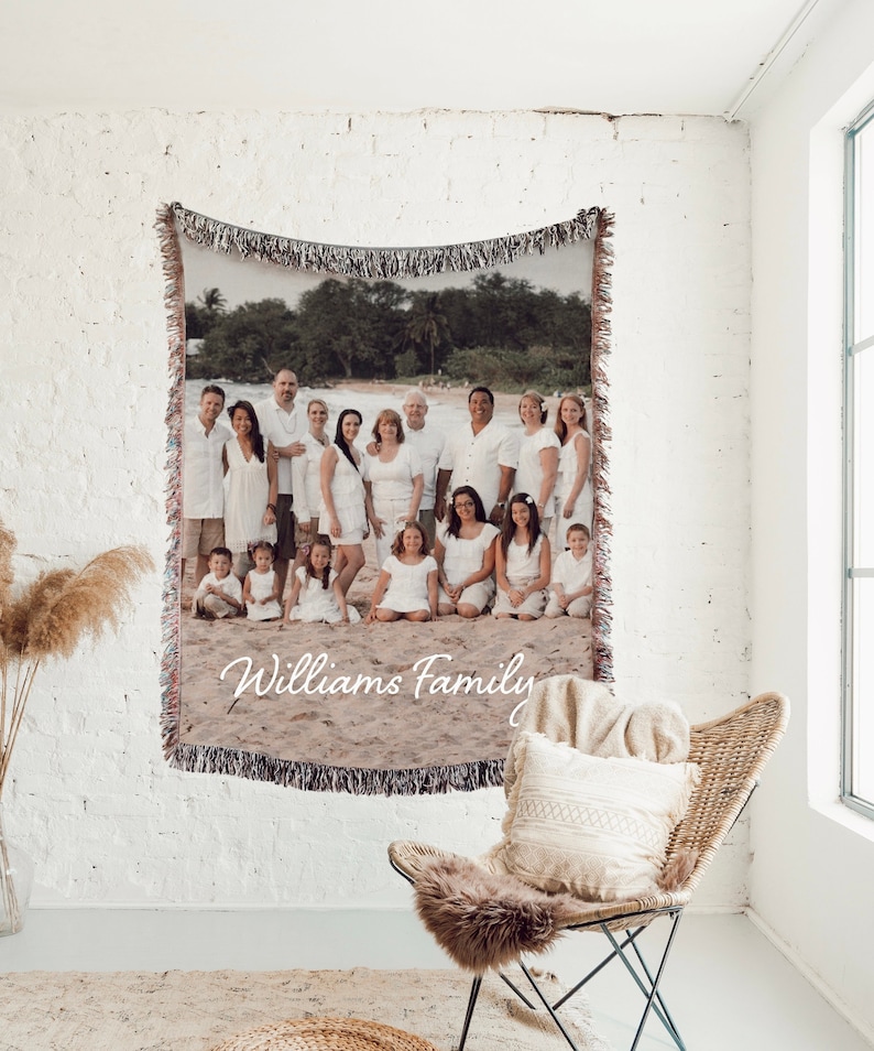May include: A woven tapestry blanket featuring a family portrait on a beach, with the words "Williams Family" in cursive. The blanket is fringed and hangs on a white brick wall. A wicker chair with a pillow and fur throw is in the foreground.