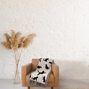 Pode incluir: Uma poltrona de couro marrom com uma manta de arremesso com um padr&atilde;o de gato preto contra um fundo de parede de tijolo branco. Um vaso com grama de pampas seca est&aacute; em primeiro plano.
