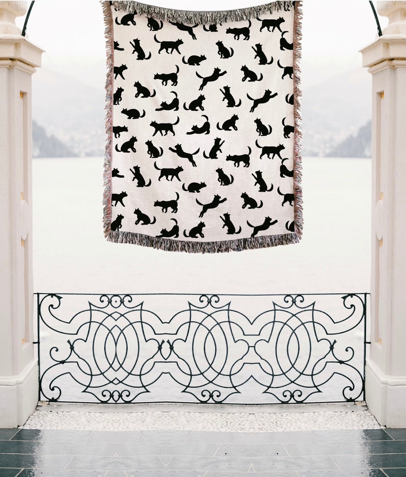 Pode incluir: Uma manta branca com um padr&atilde;o de silhuetas de gatos pretos. A manta est&aacute; pendurada em uma balaustrada branca com um design de metal preto.