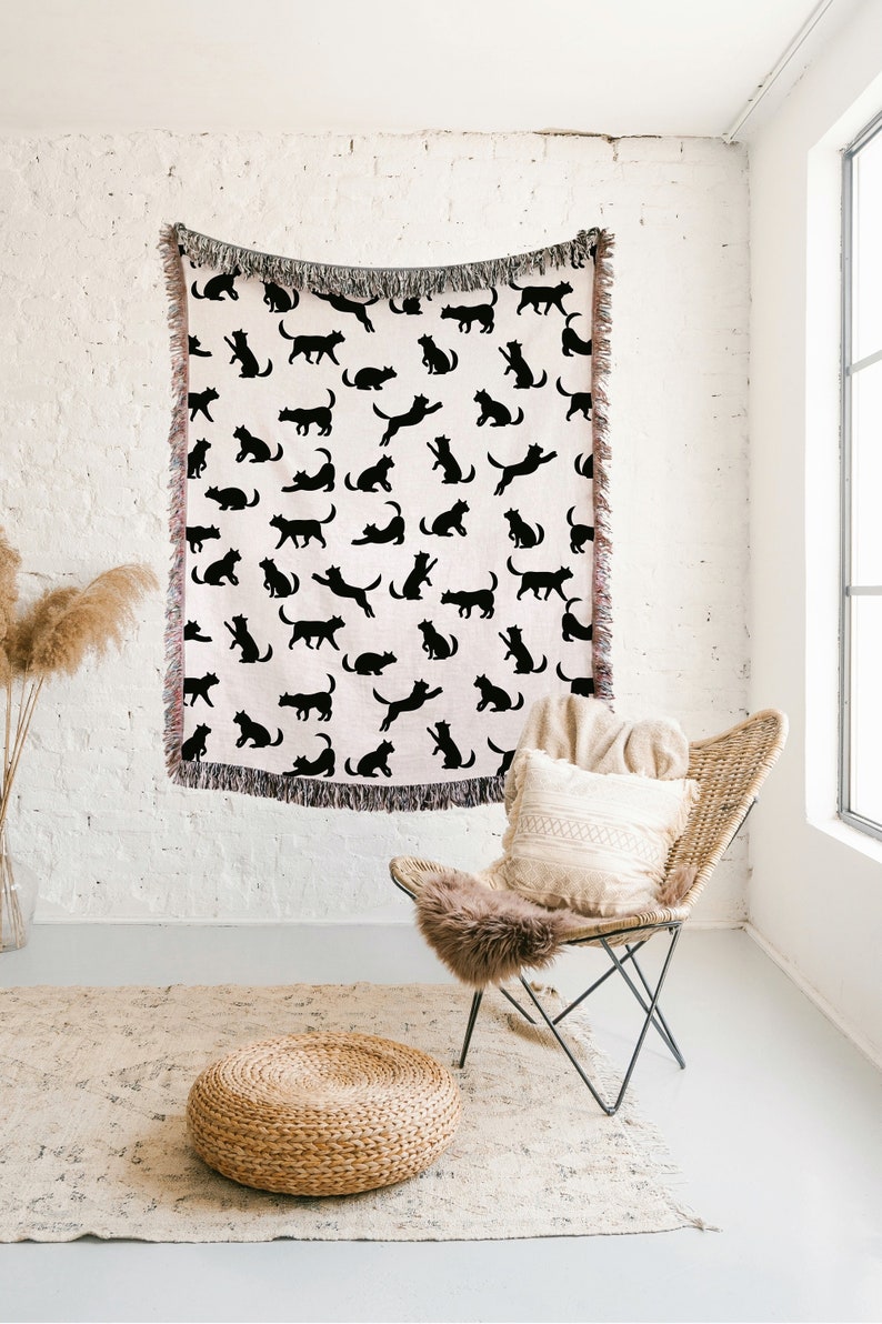 Pode incluir: Uma manta de arremesso rosa com um padr&atilde;o de gato preto. A manta est&aacute; pendurada em uma parede de tijolo branco. Uma cadeira de vime com uma almofada branca e uma manta de pele marrom est&aacute; em primeiro plano. Um pufe tecido est&aacute; em um tapete bege.