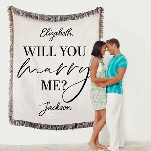 May include: A white woven throw blanket with the text "Elizabeth, WILL YOU marry ME? - Jackson" in black. A couple is standing in front of the blanket, embracing.