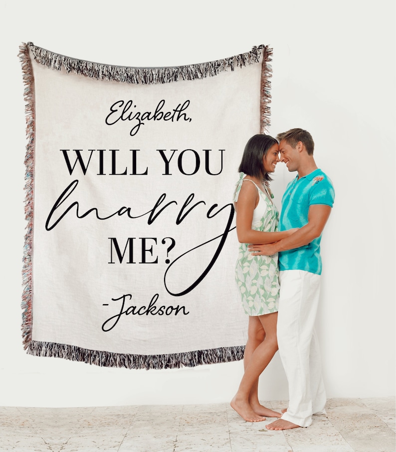 May include: A white woven throw blanket with the text "Elizabeth, WILL YOU marry ME? - Jackson" in black. A couple is standing in front of the blanket, embracing.