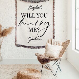 May include: A white throw blanket with the text "Elizabeth, Will You Marry Me? - Jackson" in black script font. The blanket is draped over a white brick wall and a wicker chair with a white throw pillow is in the foreground.