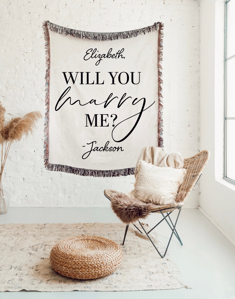 May include: A white throw blanket with the text "Elizabeth, Will You Marry Me? - Jackson" in black script font. The blanket is draped over a white brick wall and a wicker chair with a white throw pillow is in the foreground.