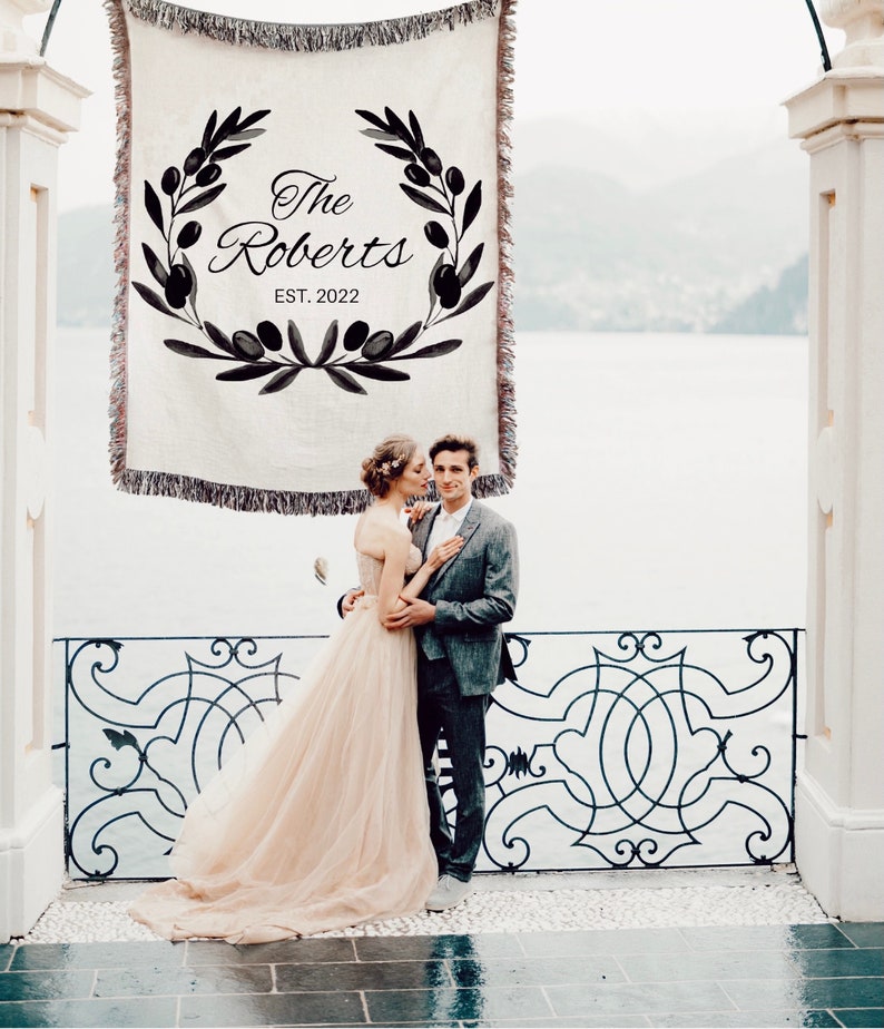 May include: A couple pose for a wedding photo in front of a white tapestry with a black olive branch wreath and the text "The Roberts EST. 2022".