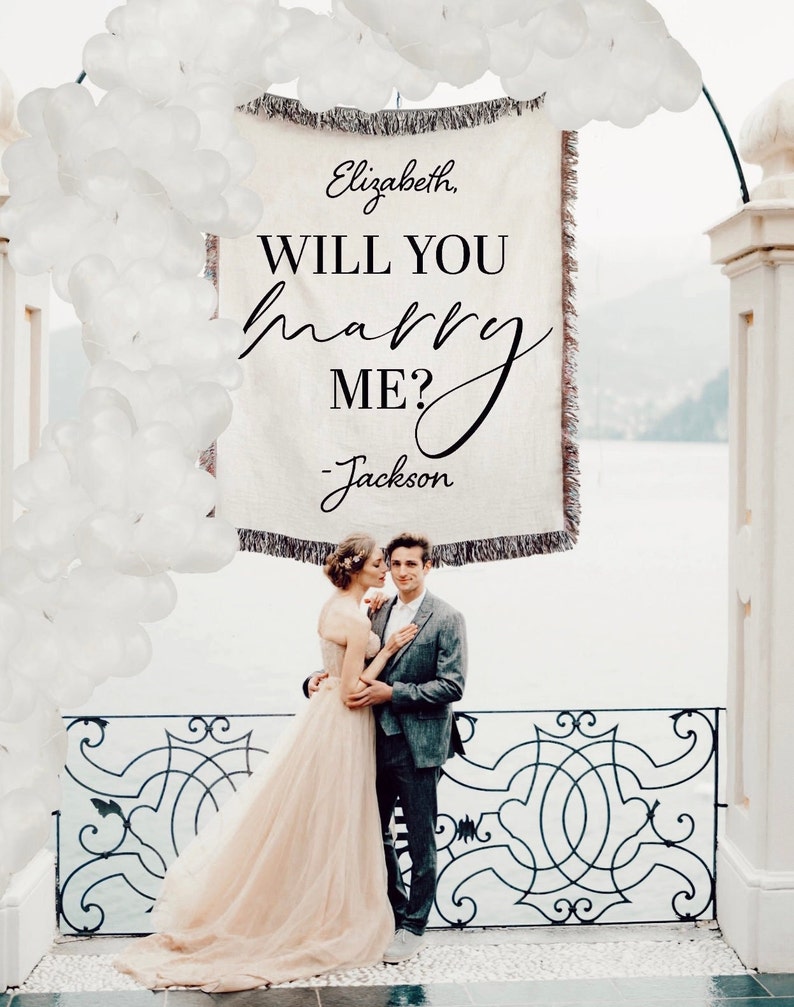 May include: A couple is standing in front of a white banner with the text "Elizabeth, WILL YOU marry ME? - Jackson". The banner is decorated with white balloons and is hanging from a white archway. The couple is standing on a balcony with a wrought iron railing.