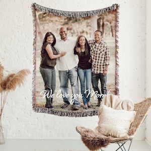 May include: A woven tapestry with a family portrait, fringed edges, and the text "We love you Mom" displayed on a white brick wall. The tapestry features a family of four, with a neutral color palette.