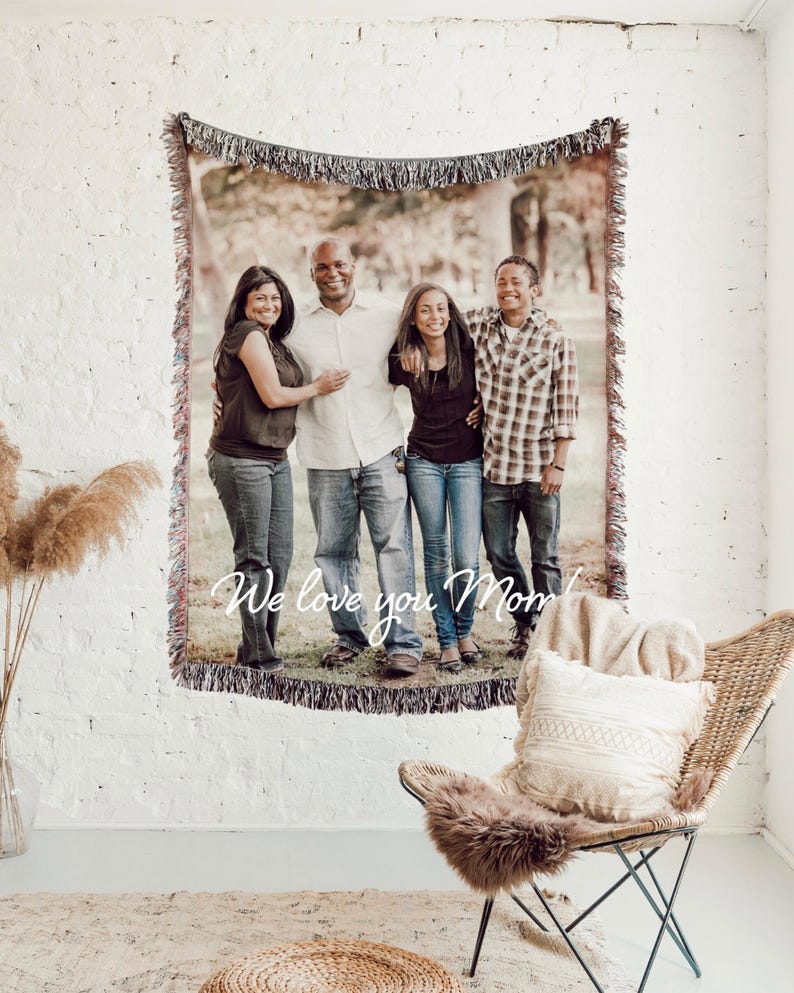 May include: A woven tapestry with a family portrait, fringed edges, and the text "We love you Mom" displayed on a white brick wall. The tapestry features a family of four, with a neutral color palette.