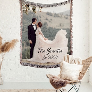 May include: Woven tapestry featuring a wedding photo of a couple in formal attire, set against a mountain backdrop. The tapestry includes the text "The Smiths Est. 2024" in elegant script, with fringed edges.