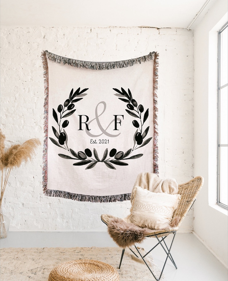 Peut inclure: Une couverture en tissu tiss&eacute; avec un motif de branche d'olivier noir et blanc et les initiales "R&F" en gris avec le texte "Est. 2021" en dessous. La couverture est accroch&eacute;e &agrave; un mur en briques blanches.