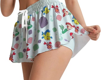 May include: A light blue athletic shorts with a white elastic waistband and a layered ruffle hem. The shorts feature a colorful print of Ariel, Flounder, Sebastian, and other characters from the Disney movie The Little Mermaid.