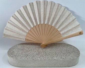 May include: A white hand fan with a wooden frame and a gold leaf design on the handle. The fan is resting on a gray, speckled surface.