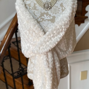 May include: A cream-colored faux fur shawl draped over a mannequin. The shawl has a textured, rose-like pattern. A multi-strand pearl necklace with a floral brooch adorns the mannequin's neck. The background features a staircase.