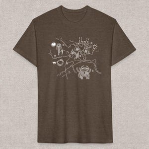 Puede incluir: Una camiseta marrón con un dibujo lineal blanco de antiguos petroglifos. El diseño incluye figuras, símbolos y un diseño similar a un mapa. La camiseta está hecha de una tela suave y texturizada.