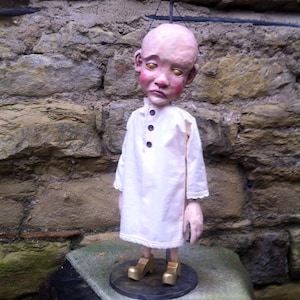 May include: A wooden doll with a bald head and large eyes wearing a white dress with lace trim and gold shoes. The doll is standing on a wooden platform with a brown rug.