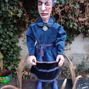 May include: A handmade marionette puppet of a woman with a stern expression. The puppet wears a blue dress with a black skirt, a jeweled brooch, and blue shoes. The puppet's face is painted with detailed features, and it has a carved wooden head.