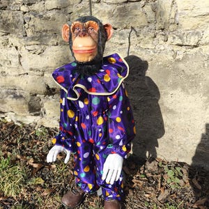 May include: A whimsical monkey doll dressed in a purple satin clown suit with colorful polka dots. The doll has a detailed monkey face, white gloves, brown shoes, and a ruffled collar. It stands on grass in front of a stone wall.