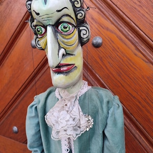 May include: A hand-painted wooden puppet with a green and white outfit, a white lace collar, and a large, expressive face with a long nose.